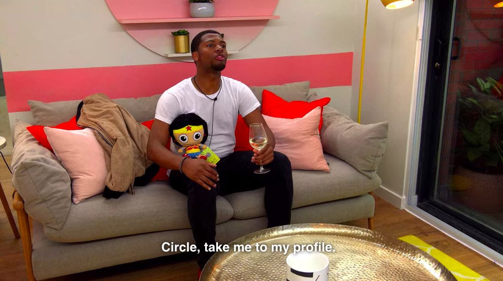 A character on the reality show The Circle holds a stuffed doll, a glass of wine, and asks to see his profile