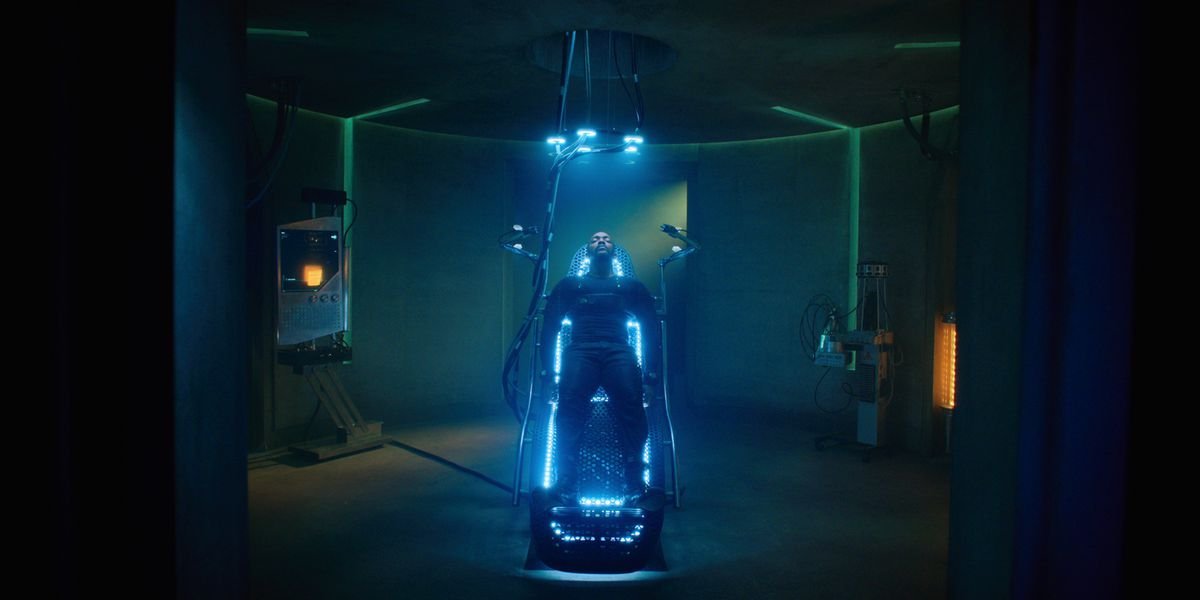 Anthony Mackie lies on a surgical table, wearing a full-body black jumpsuit, lit by blue neon lights built into the table and hanging above him.