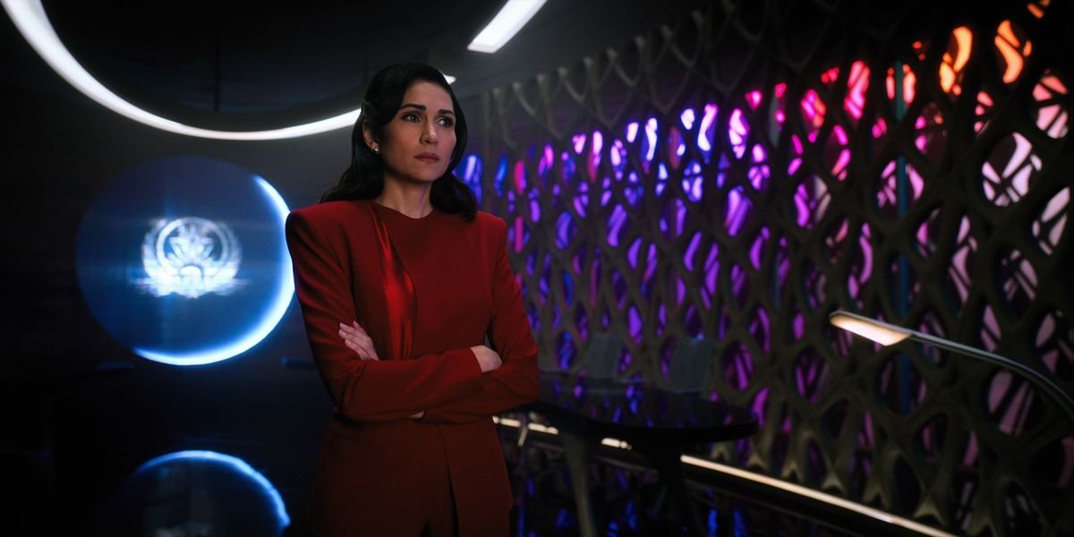 A woman in a blood-red suit stands in a dark room with a sigil glowing in the window behind her, and a bank of multicolored lights shining through cutouts in the wall next to her.