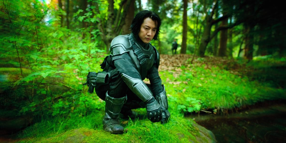 An Asian man in matte grey body armor squats by a small body of water in an outdoor setting where the vegetation glows a vivid bright green.