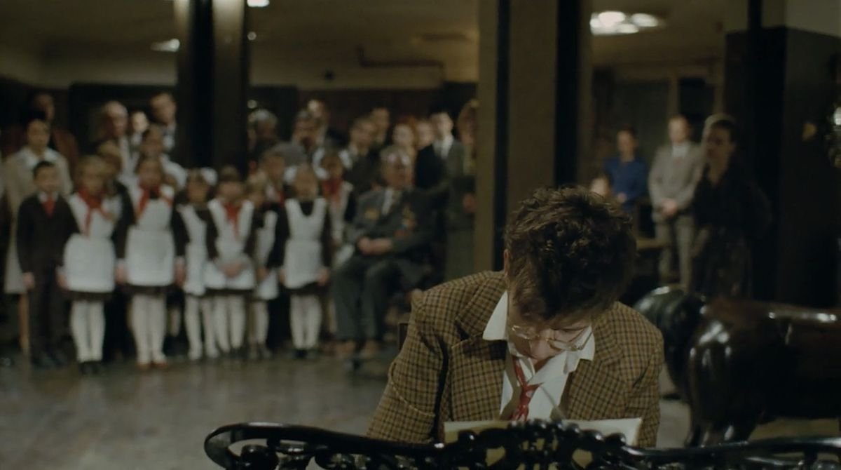 A bespectacled, tousle-haired man in a brown suit jacket and red tie plays the piano in the foreground, as in the background, a crowd of standing people of various ages watch him.