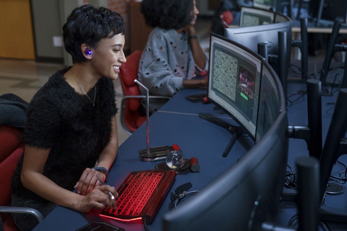 Andy Allo sits at a curved bank of computer monitors with her hands on a bright red keyboard, smiling as she looks at something onscreen.