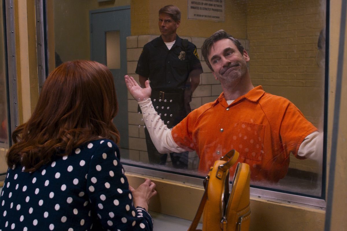 Jon Hamm as the Reverend in Unbreakable Kimmy Schmidt wears an orange prison jumpsuit and shrugs dismissively at Kimmy from the other side of a prison visitation window.