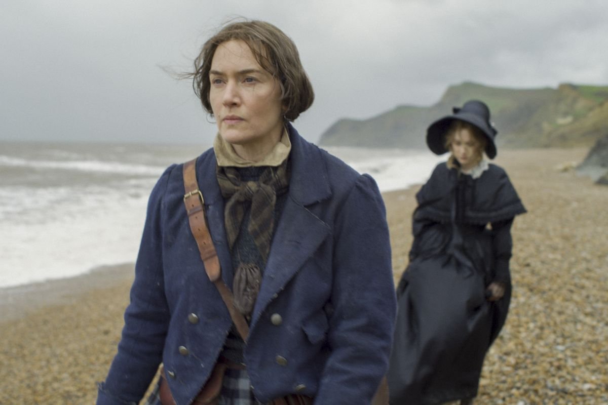 Kate Winslet walks ahead of Saoirse Ronan on a windblown beach in Ammonite