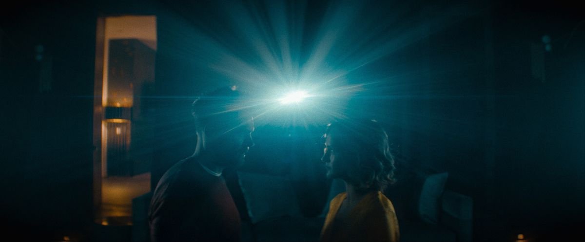 Joel McHale and Kerry Bishé face each other in a dark room with a single very bright light like a movie projector behind them