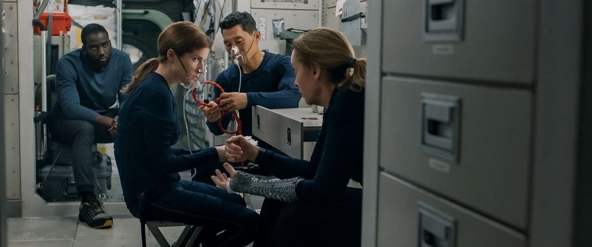 The cast of Stowaway in their galley, with Anna Kendrick and Daniel Dae Kim wearing oxygen masks, Toni Collette sitting close to them, and Shamier Anderson sitting in the background