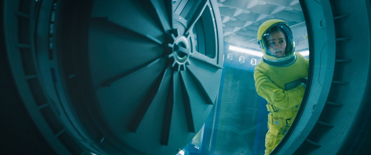 A nervous-looking teenage girl in a yellow spacesuit, peering out through a huge round hatch in Voyagers