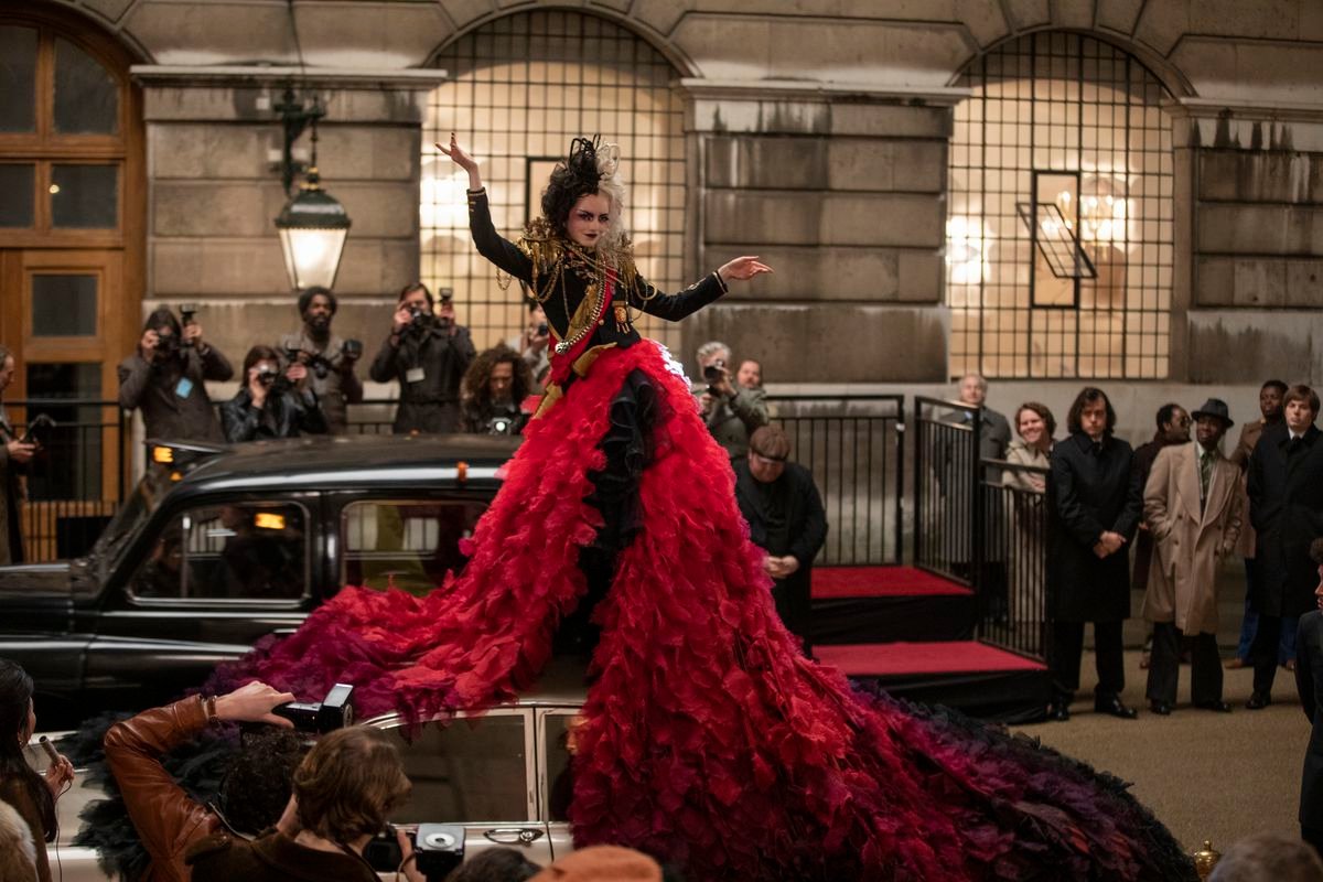 cruella in a tumbling red gown standing on top of a car