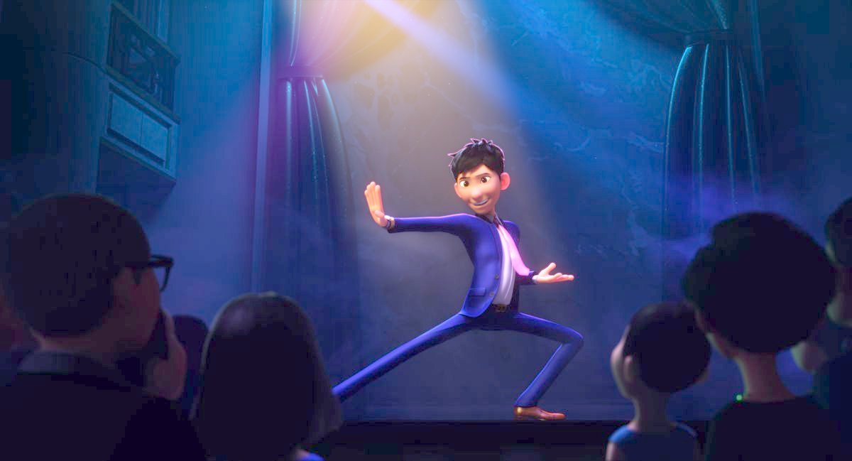 Din, in a blue suit and pink tie, poses in a dramatic kung-fu-style pose onstage in front of an audience in Wish Dragon