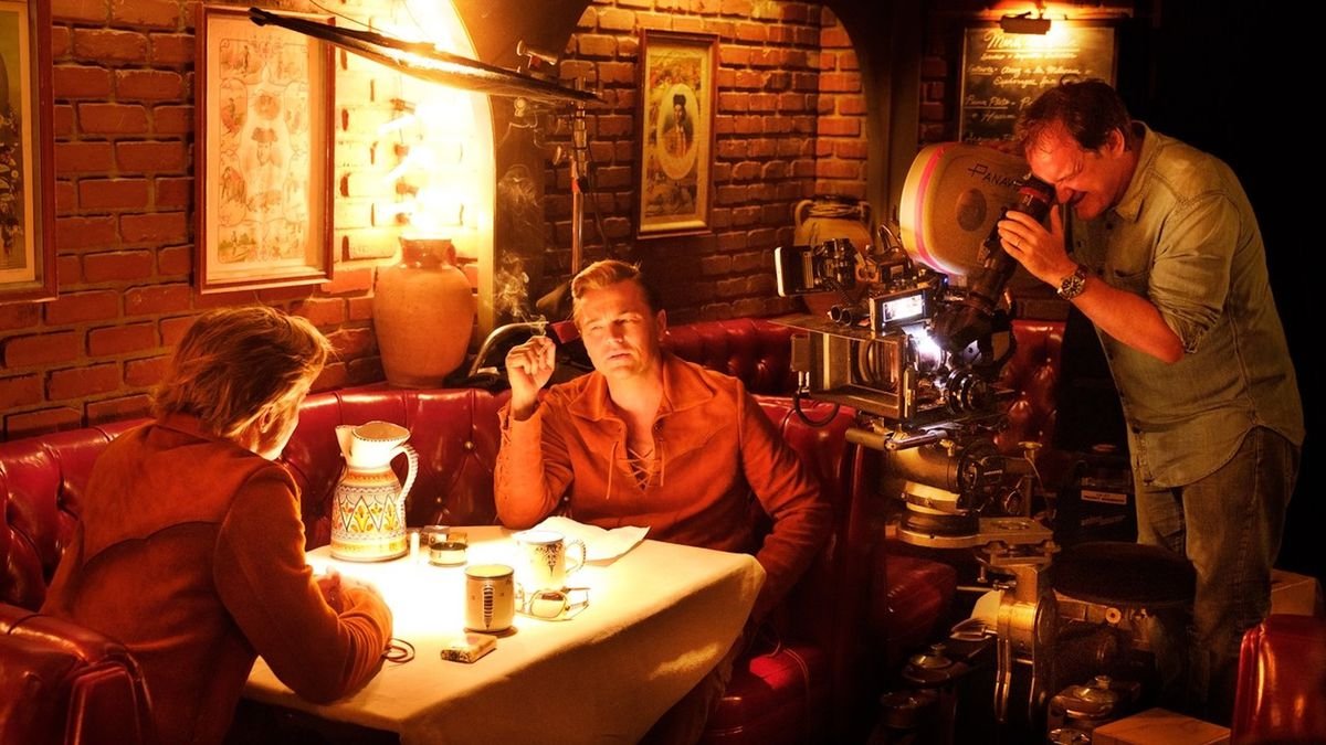 Brad Pitt and Leonardo DiCaprio sit facing each other in a restaurant booth as Quentin Tarantino watches from behind the camera filming Once Upon a Time ... in Hollywood