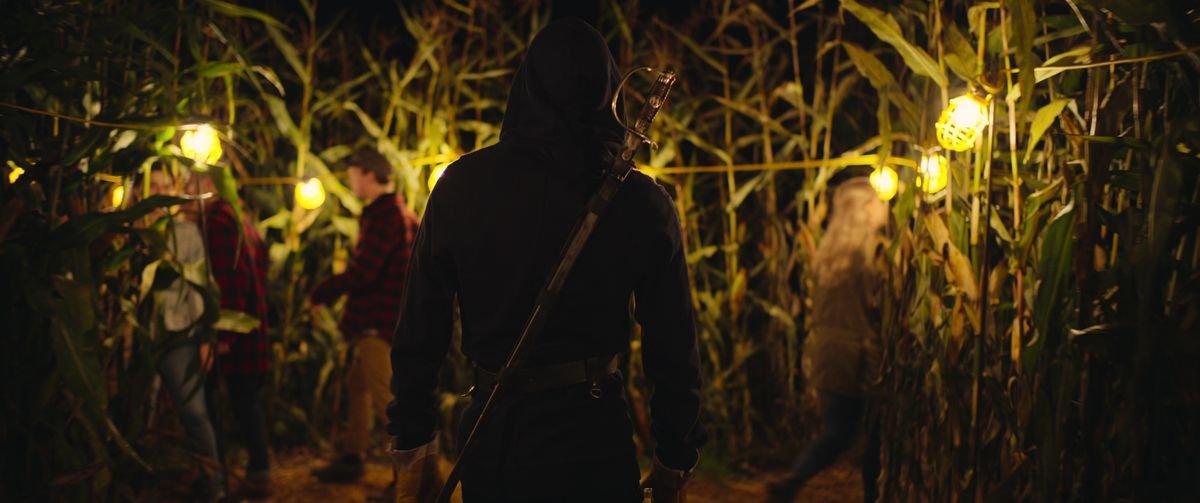 A hooded figure with a sword across their back stands in a cornfield, back to the camera, as other out-of-focus people enter the corn in There’s Someone Inside Your House