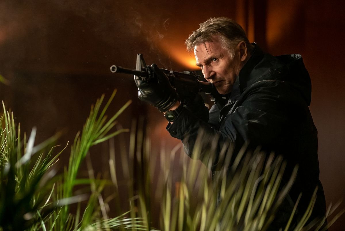 Liam Neeson stalks through tall grass with an assault rifle in Memory