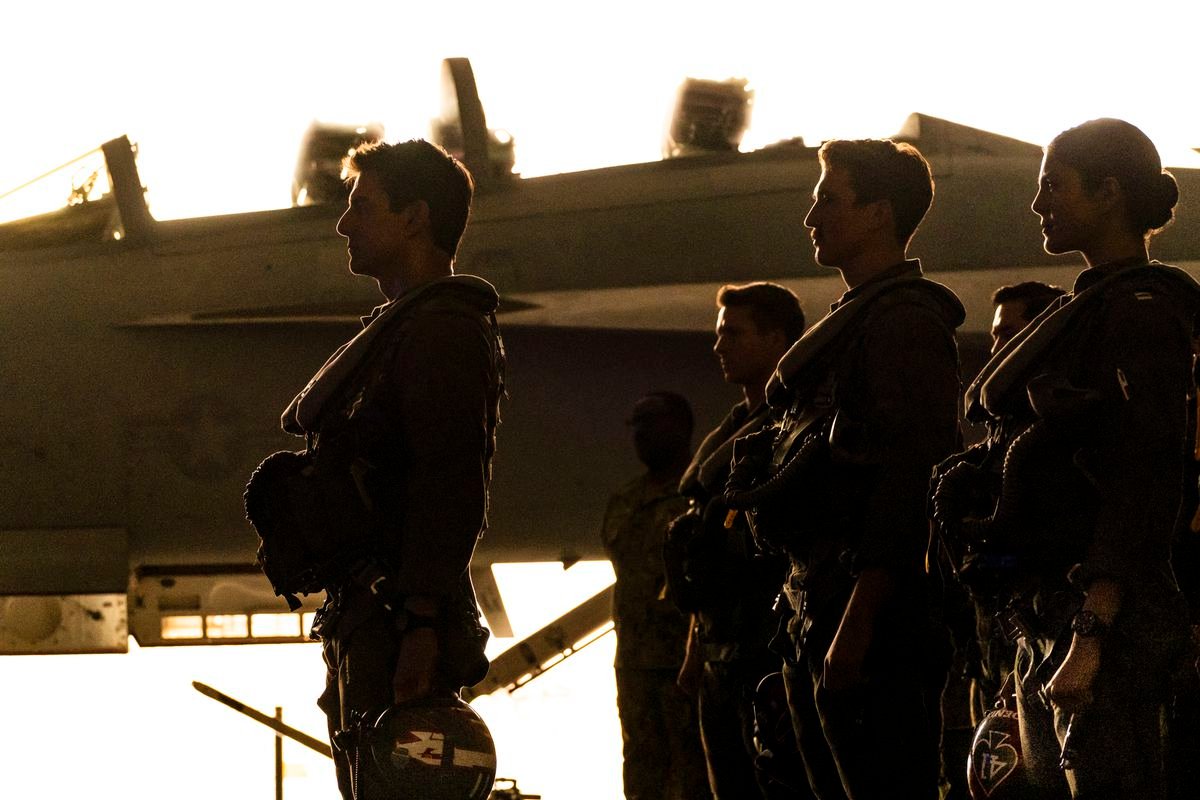 Tom Cruise, Miles Teller, Monica Barbaro and other young pilots stand in silhouette with a jet in the background