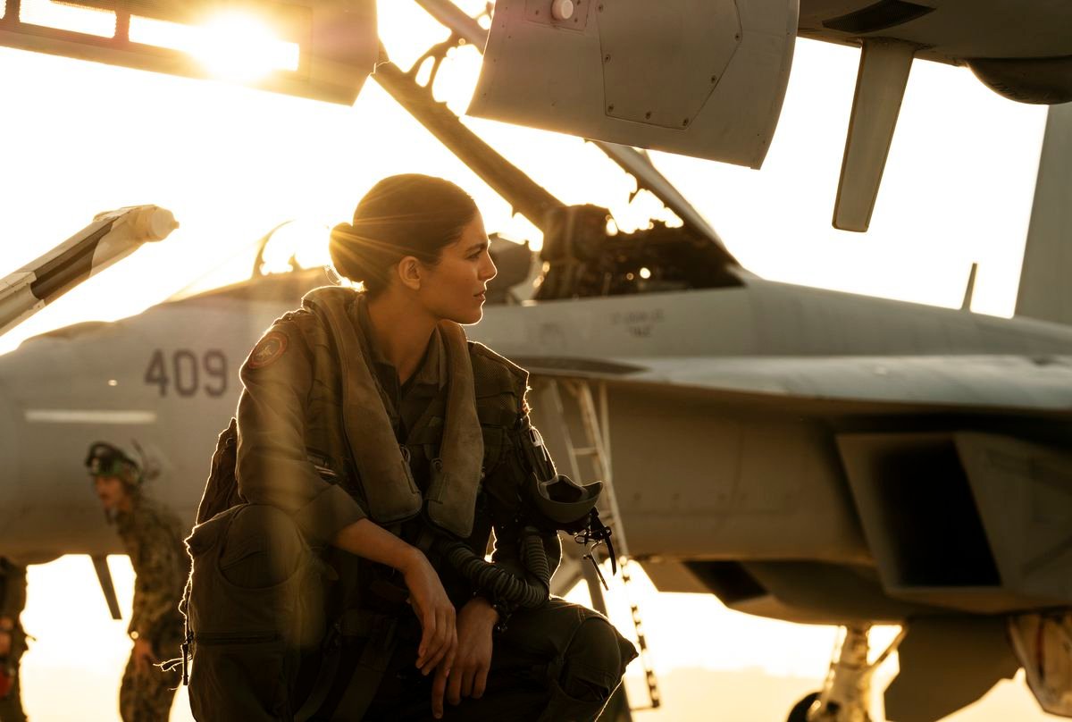 Phoenix (Monica Barbaro) squats near some fighter jets with the sun behind her