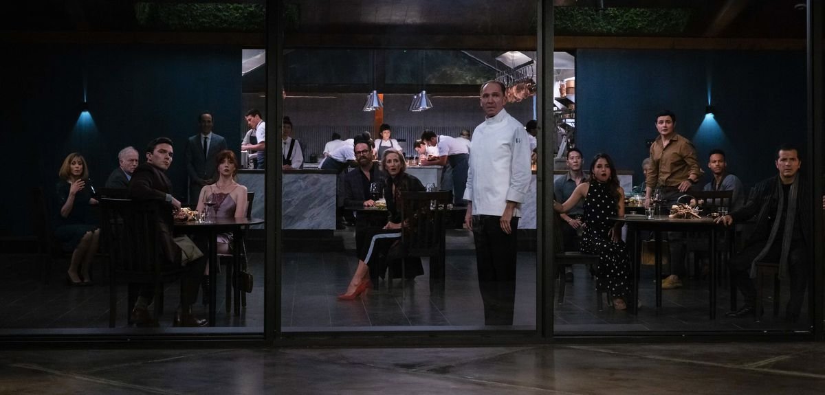 Chef Slowik stands in a large windowed dining area surrounded by restaurant patrons who are all turned toward the windows, looking in shock at something off screen