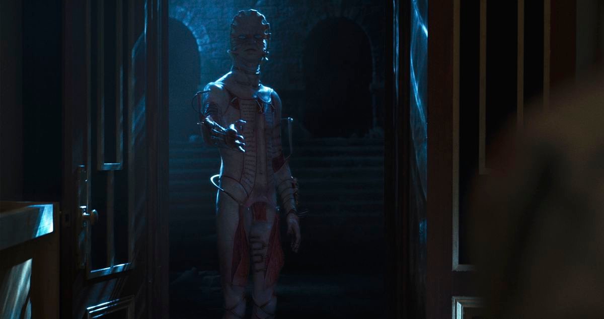 A cenobite with flayed skin and a flat head enters from the darkness in a mansion room