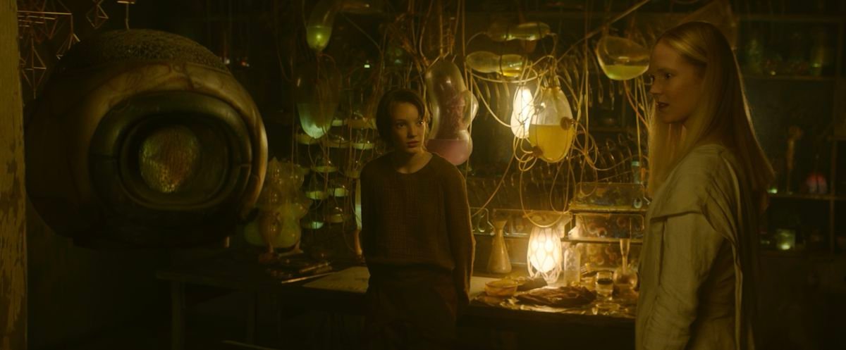 Vesper (Raffiella Chapman) and Camellia (Rosy McEwen) stand in Vesper’s dark, crowded lab in Vesper