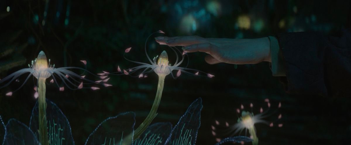 Vesper (Raffiella Chapman) holds her hand over a delicate, glowing flower that reaches its tendrils toward her in Vesper