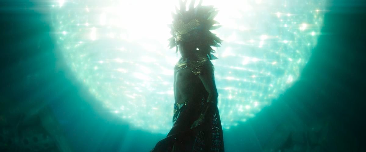 A man in an elaborate feather headdress (played by Tenoch Huerta) and cape swims in front of a gigantic glowing sphere in Black Panther: Wakanda Forever.