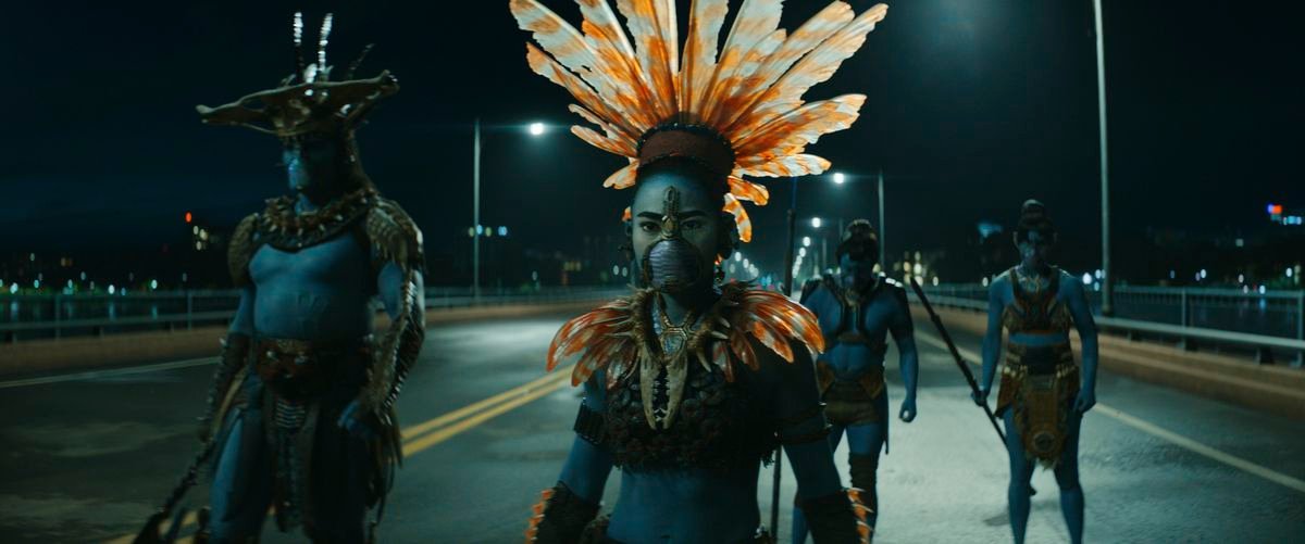 A group of blue-skinned Talokan warriors stand in the middle of a bridge at night, one of them wearing an elaborate orange feathered helmet, in Black Panther: Wakanda Forever.