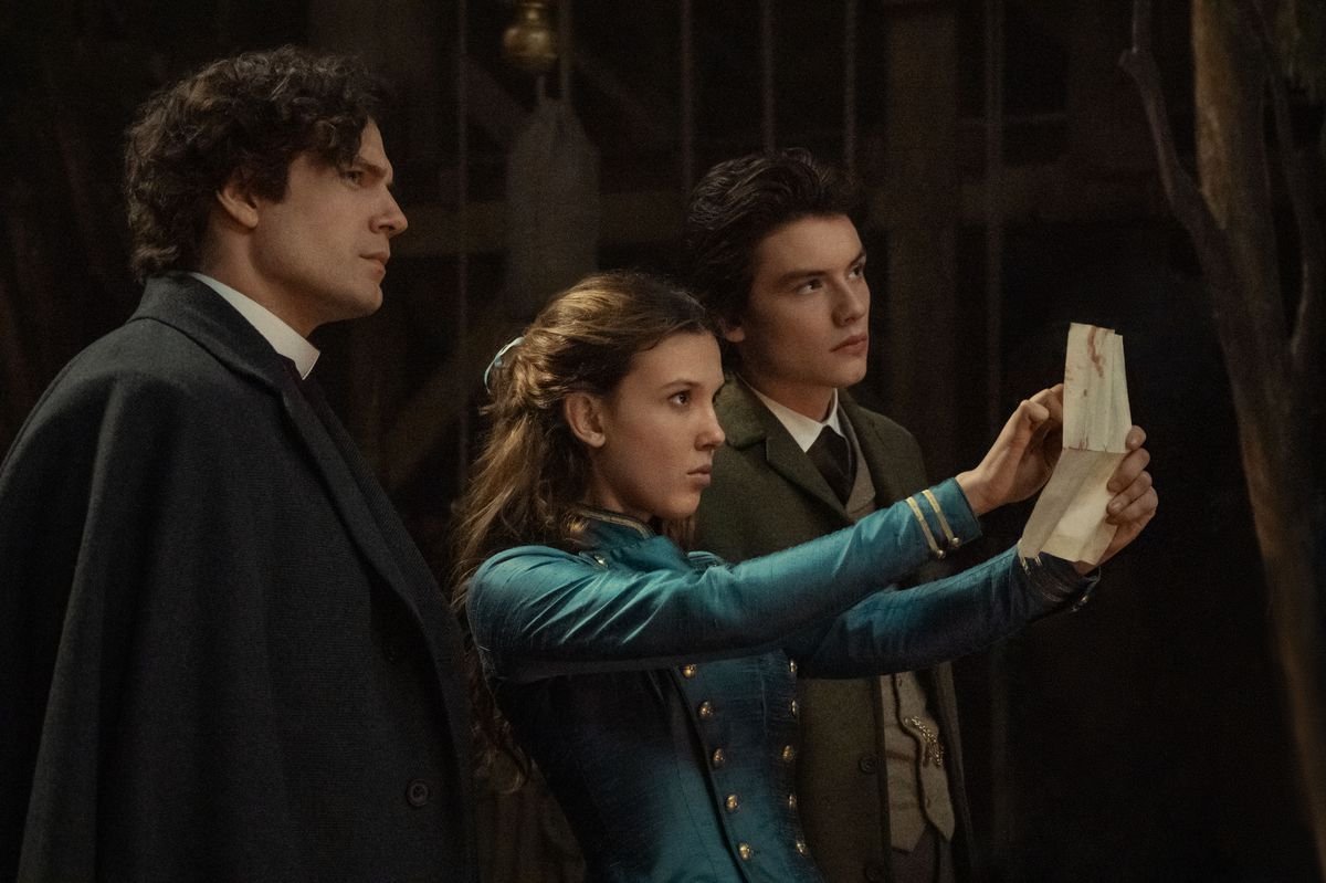 Henry Cavill as Sherlock Holmes, Millie Bobby Brown as Enola Holmes, Louis Partridge as Tewkesbury in Enola Holmes 2. Enola holds up a piece of paper and points at it while the others look on.