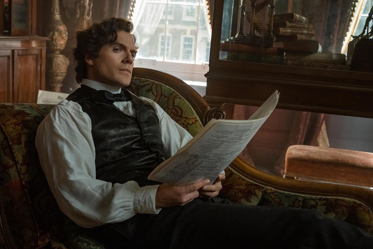 henry cavill as sherlock holmes. he sits on a chaise lounge, peering at some papers in his hands