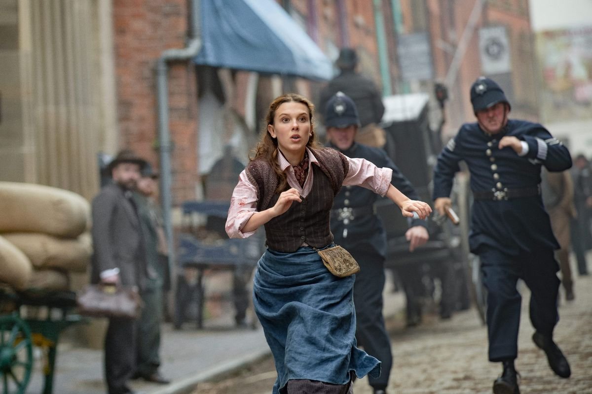 millie bobby brown as enola holmes. she runs through the london streets, two police officers right on her tail