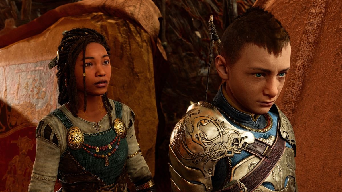 Angrboda, a Black teenage girl wearing overalls with her hair in dreadlocks, stands behind Atreus, a white teenage boy in gold and blue armor, looking at him in God of War Ragnarök