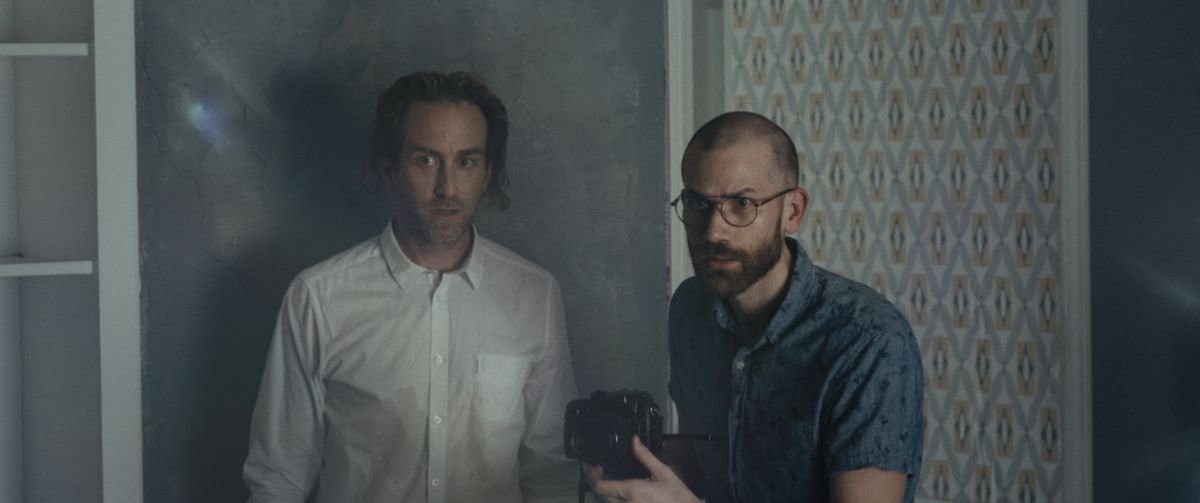 Levi (Justin Benson) and John (Aaron Moorhead) stand against an elaborately patterned wall, aiming a camera at something offscreen that they’re staring at distrustfully in Something In the Dirt