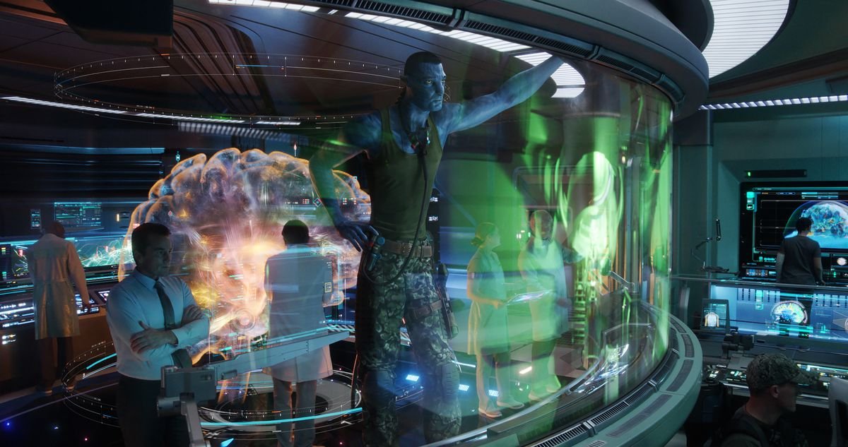 The Na’vi form of Col. Quaritch (Stephen Lang) stands in a command center surrounded by humans and looks at an elaborate VR display in Avatar: The Way of Water.