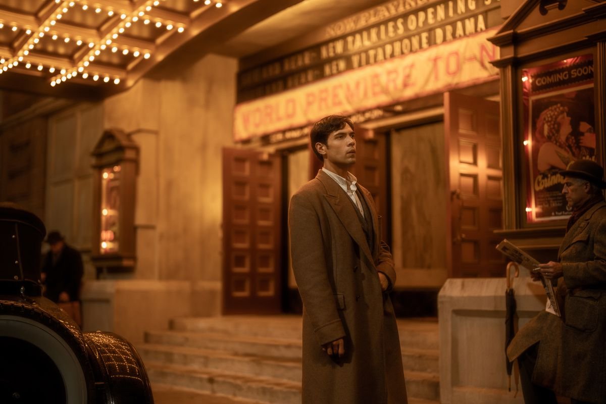 Manny stands in a trench coat under the awning of a movie palace, in front of the marquee posters of classic Hollywood in the film Babylon