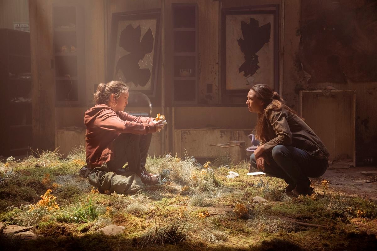 Ellie and Tess crouch facing each other in the grass that has overtaken a decrepit building as light shines in from above in the HBO series The Last of Us.