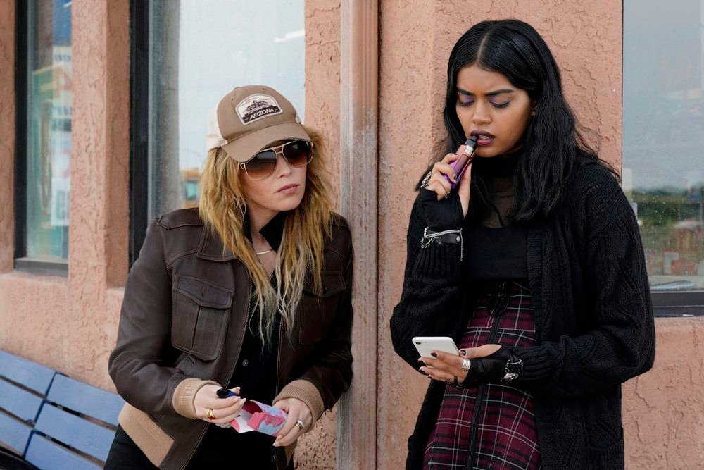 Natasha Lyonne as Charlie Cale, wearing a trucker hat and big sunglasses, leans in as Sara (Megan Suri ) nibbles on her vape and looks at her phone in Poker Face