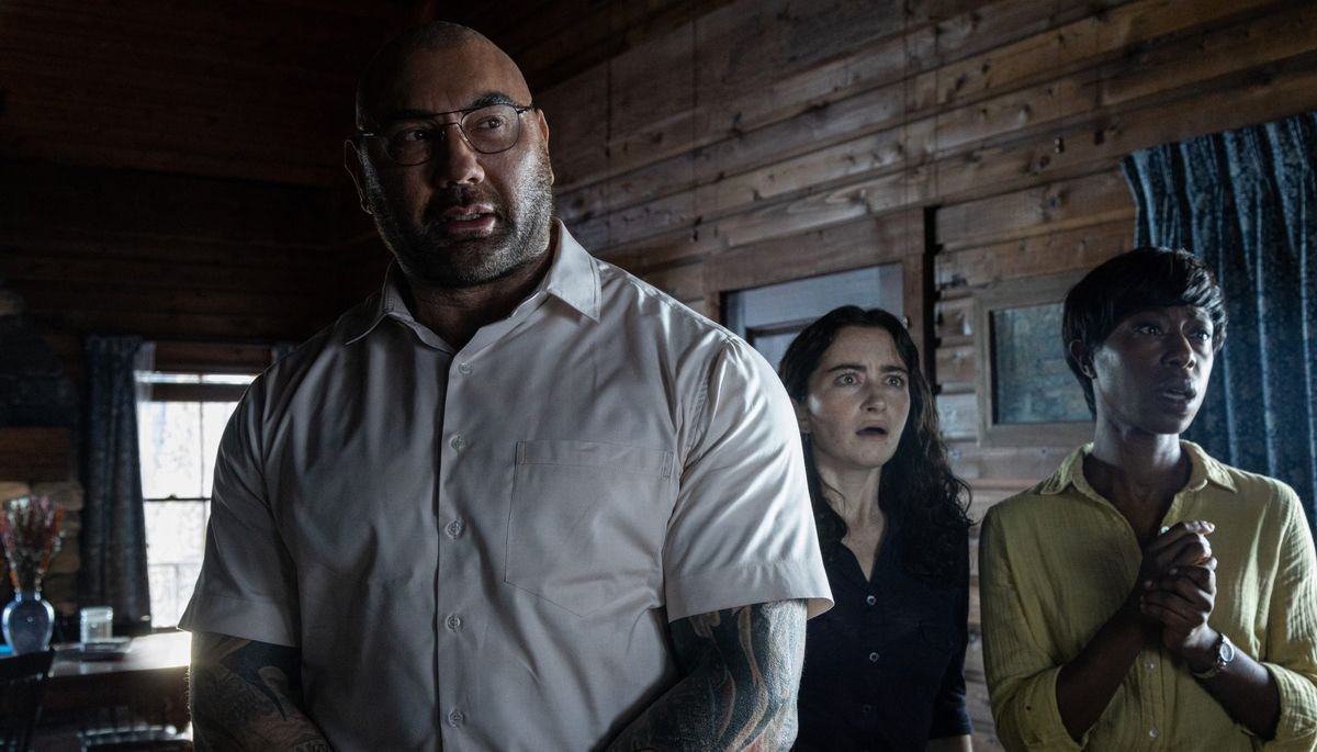 Home invader Leonard (Dave Bautista) stands nervously in front of companions Sabrina (Nikki Amuka-Bird) and Ardiane (Abby Quinn) as they all stare at something offscreen in Knock at the Cabin