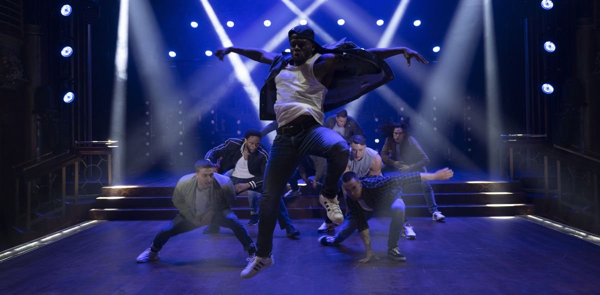 A group of (fully clothed, sorry) male dancers crouch on a stage lit with deep-blue light and crosshatched with spotlights as one dancer leaps upward with his arms spread in Magic Mike’s Last Dance