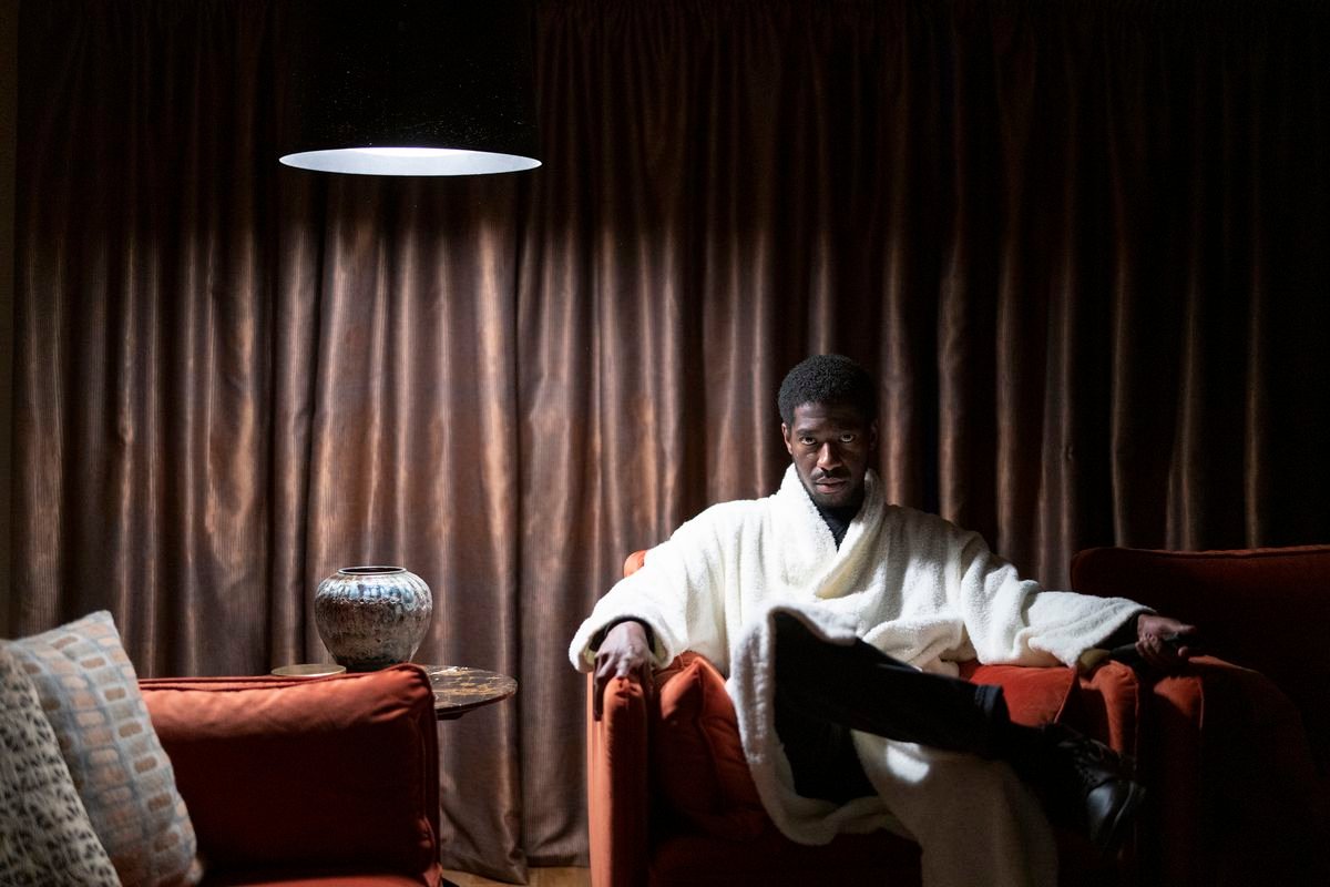 A young Black man (Jorden Myrie) in a fluffy white bathrobe sits in a plush red chair in a dark, curtained room and stares directly at the camera in a scene from Netflix’s The Strays