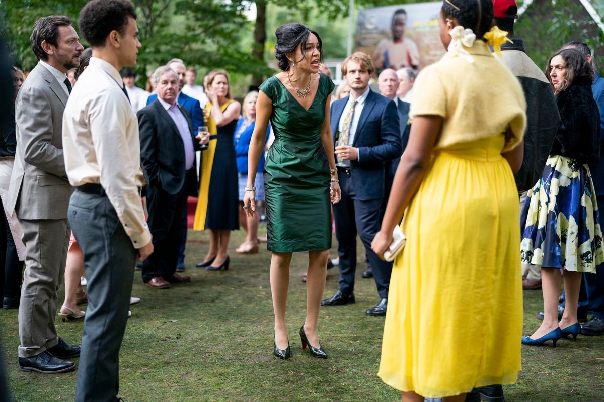 A young Black woman in a bright yellow dress stands in a crowd of people outdoors at what appears to be a garden party and yells at them in a scene from Netflix’s The Strays