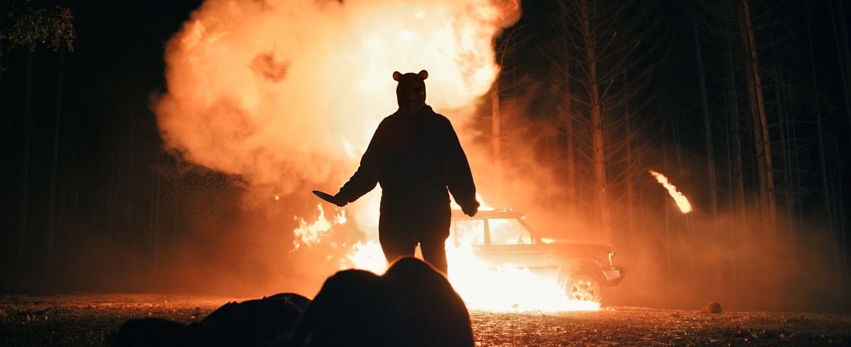 Winnie the Pooh (Craig David Dowsett) stands silhouetted against an exploding truck in the woods at night and holds up a knife as he approaches another victim in Winnie the Pooh: Blood and Honey