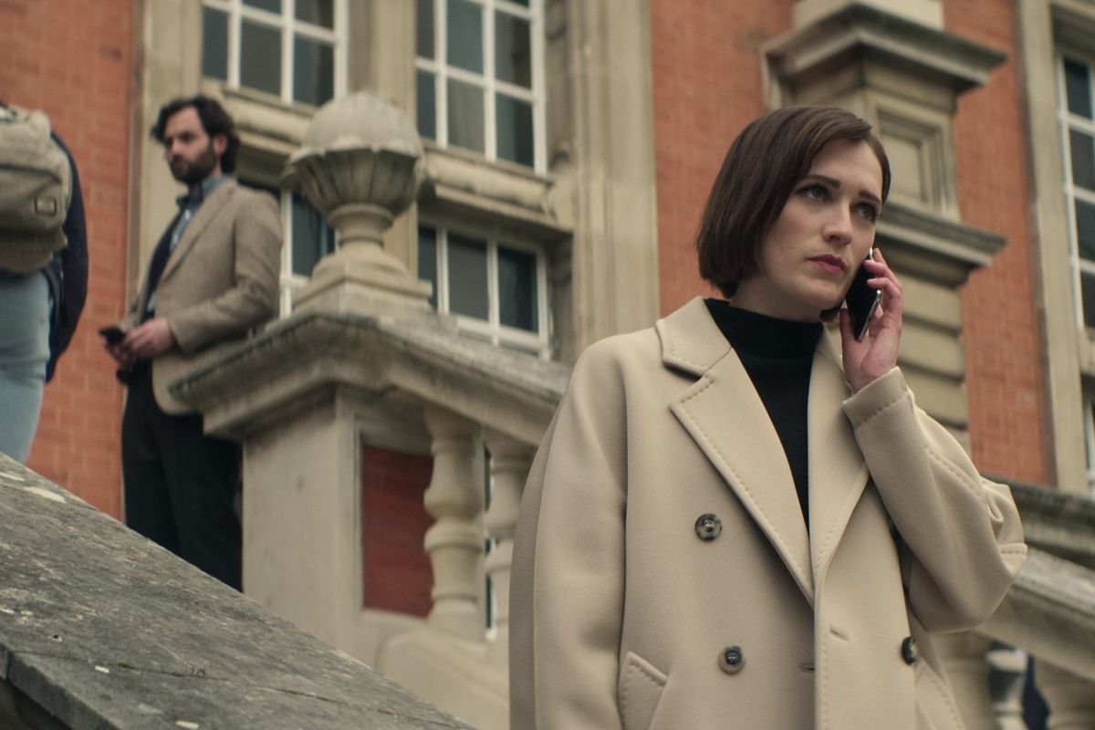 Joe lurks in the background at the top of an outdoor staircase while Kate stands at the bottom on the phone in a trench coat in a scene from season 4 of Netflix’s You.