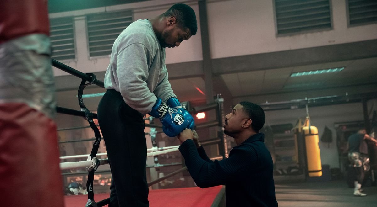 Damian (Jonathan Majors), a boxer in a grey sweatshirt, black sweatpants, and electric blue boxing gloves, leans down out of a boxing ring in a gym to bump fists with Adonis Creed (Michael B. Jordan) in Creed III