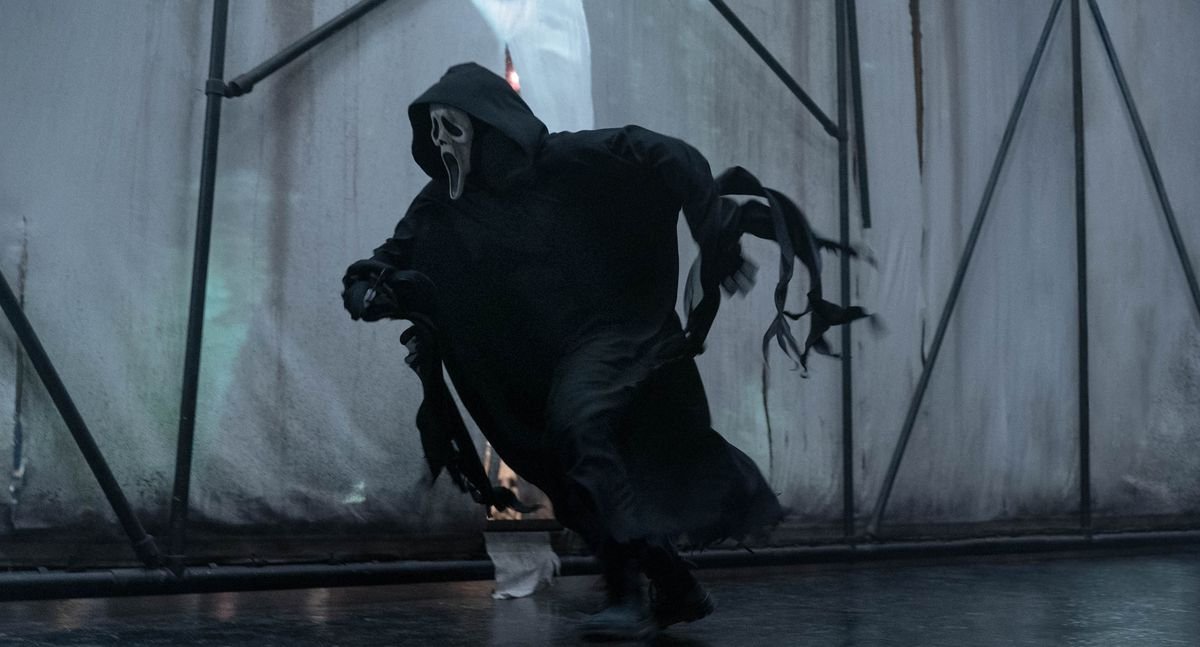 Ghostface, the black-robed, white-masked killer of the Scream franchise, runs after someone against  backdrop of white scrims held up by black scaffolding in Scream VI