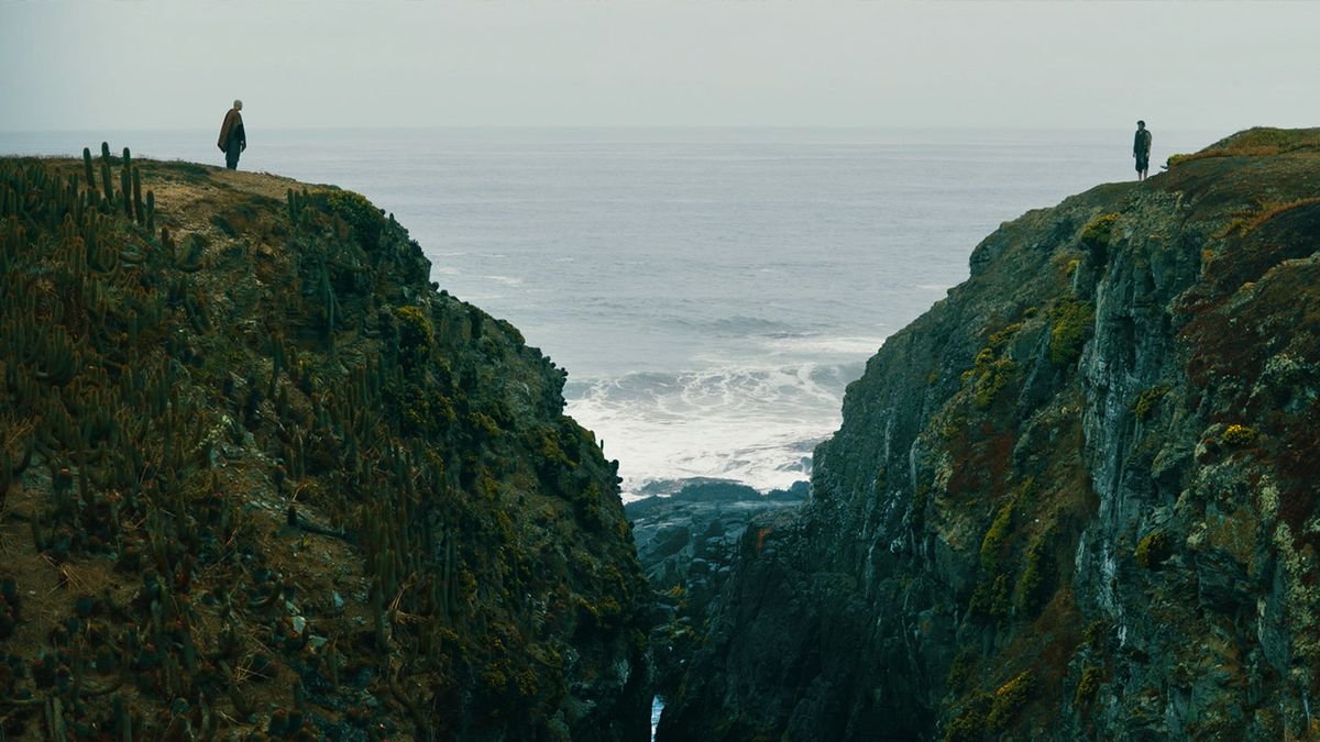 Two men stand on the top of cliffs overlooking each other, with a chasm between them and the ocean behind them, in Fist fo the Condor.