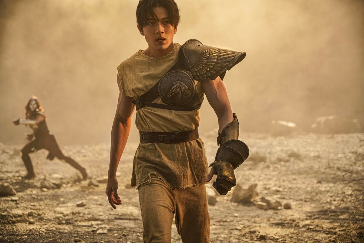 Seiya, the protagonist of the live-action 2023 Knights of the Zodiac, walks across a sandy, windblown space while wearing a brown tunic, a single metal gauntlet, and a wing-shaped brass pauldron