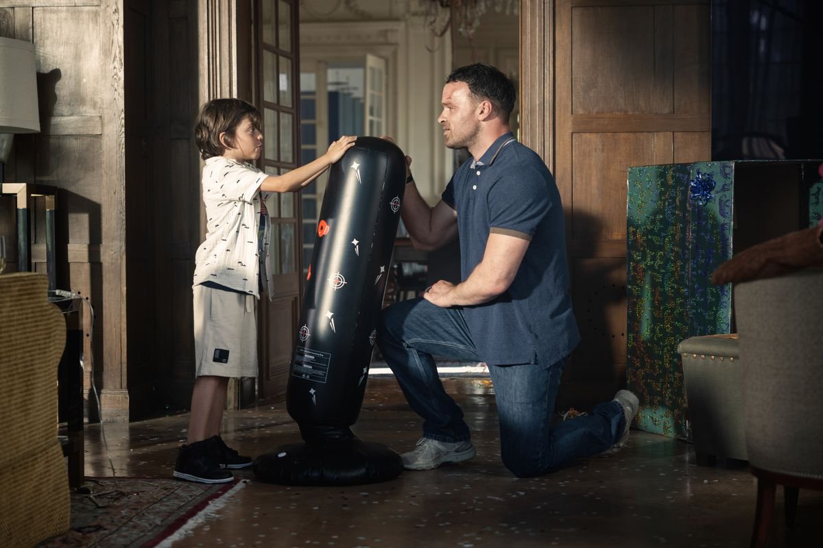 Alban Lenoir kneels down to a child’s eye level as they practice on a child’s boxing dummy in AKA.