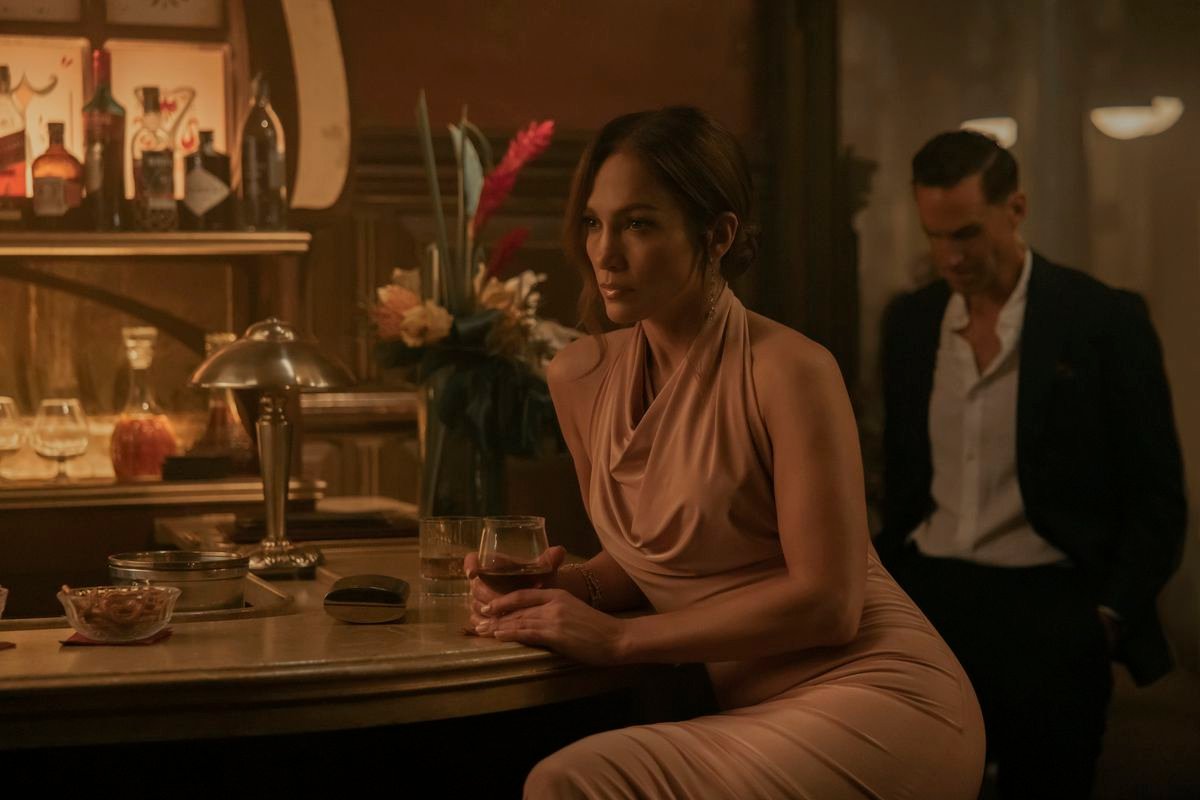 Jennifer Lopez sits at a bar with a glass of wine while wearing a nice dress in The Mother. Joseph Fiennes looms behind her, wearing a suit.