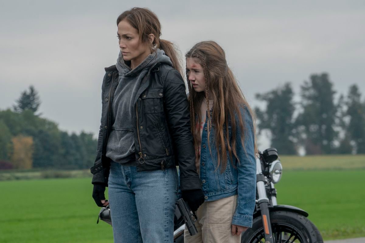 Jennifer Lopez, wearing a leather jacket, stands protectively in front of Lucy Paez, in front of a motorcycle, in The Mother.