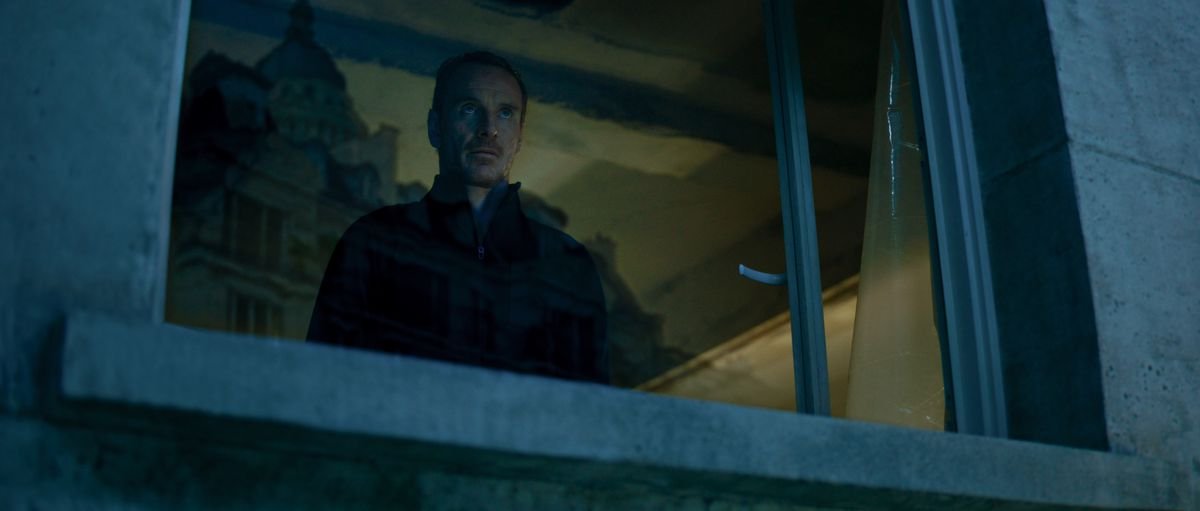 In the most David Fincher-y image possible, the unnamed Killer of The Killer stands in the window of a grimy, dimly lit room, looking out, and he’s played by Michael Fassbender, but you can barely see him because everything’s so dark and grainy