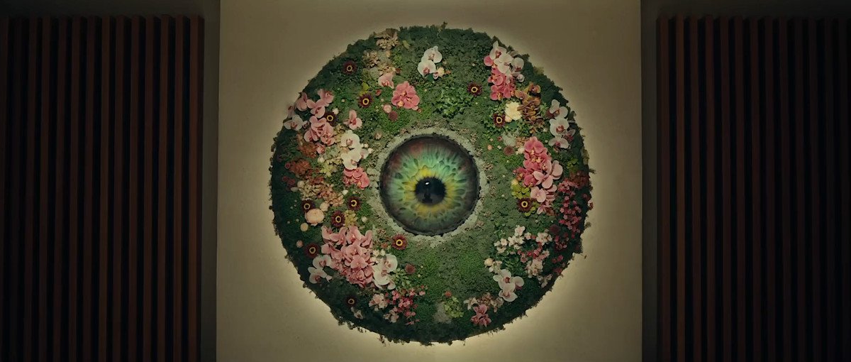 The AI therapist of The Pod Generation, a huge green eyeball built into a wall, surrounded by a circular display of green grass and pink and white flowers
