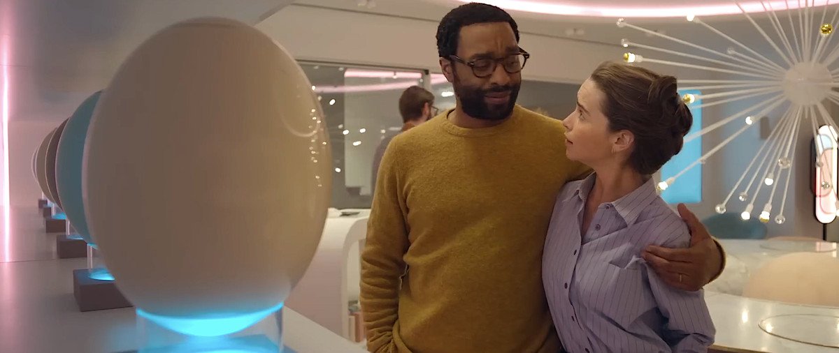 Alvy (Chiwetel Ejiofor) smiles and hugs his wife Rachel (Emilia Clarke) as they stand in a bright pastel space full of huge, shiny plastic eggs on display for sale in The Pod Generation