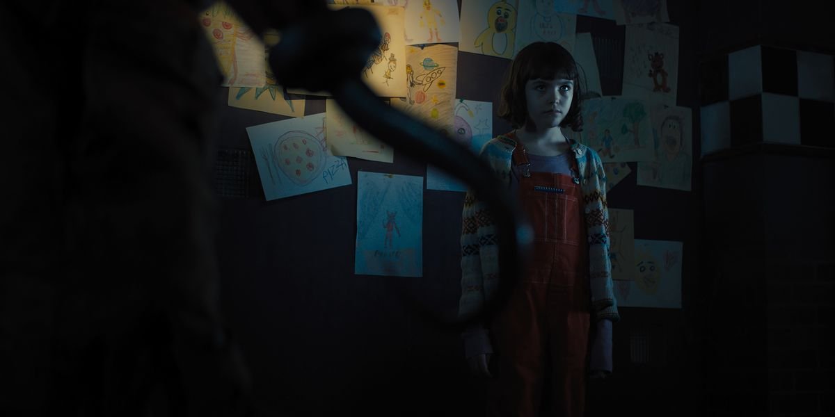 Abby (Piper Rubio), a small, pale girl with dark hair, stands in an unlit room in front of a wall covered in children’s drawings, as an unseen figure looms over her, with a hook-hand prominently in the foreground of the screen in the Five Nights at Freddy’s movie